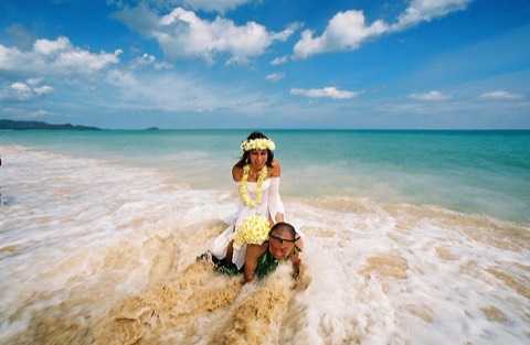 A bride in a yellow plumeria lei and haku rides on her groom in the waves on Oahu