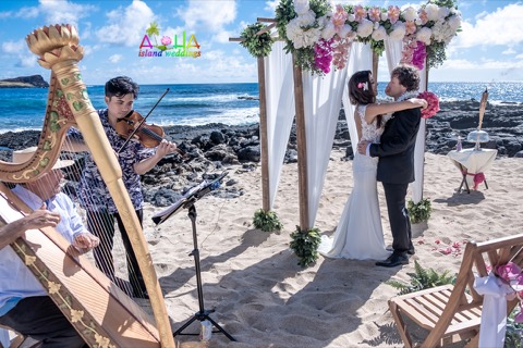 A bride and groom share their first dance under a white and pink flower arch as a violinist and harpist play