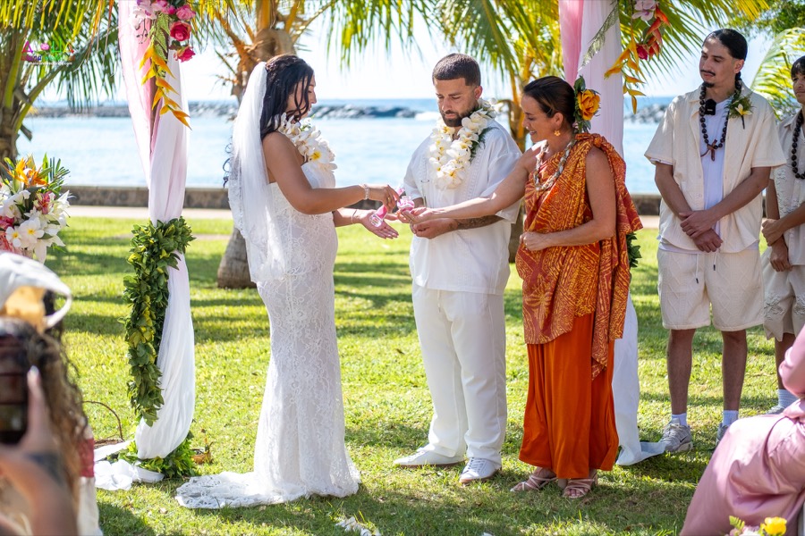 Bride and groom holding hands with the officiant during their ceremony under the flower arch by the ocean