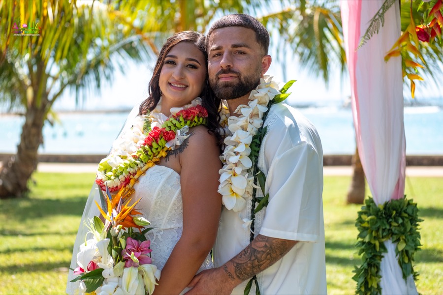 Smiling couple in flower leis embracing beside the arch with a tropical bouquet