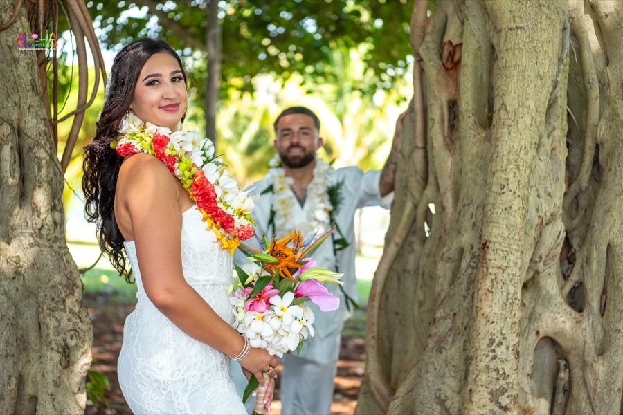 Bride holding her tropical bouquet between banyan tree roots as the groom waits behind her