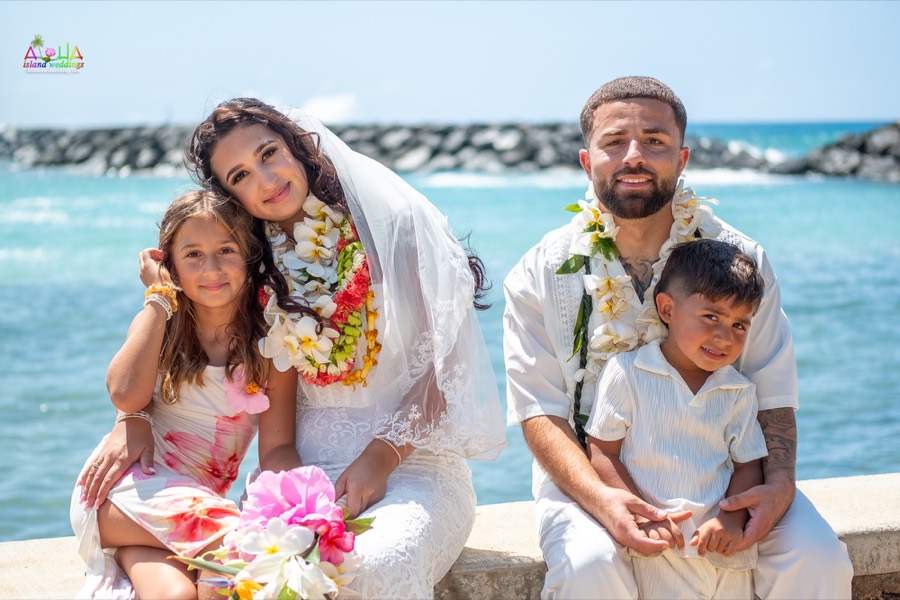 Bride, groom and their two children smiling on the seawall by the ocean