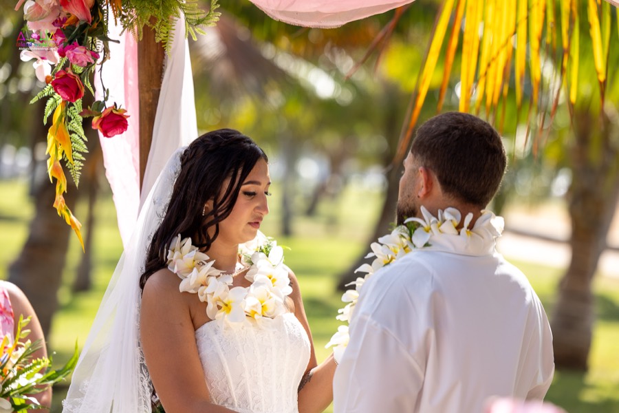Bride and groom in white plumeria leis facing each other under the pink flower arch