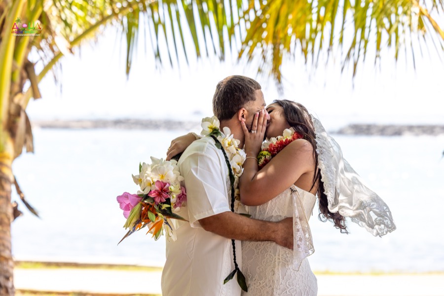 Couple kissing under the palm trees as the bride holds her tropical bouquet