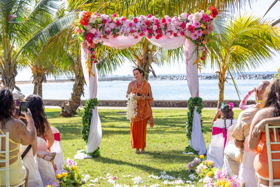 Officiant in an orange dress welcoming guests under the pink and white flower arch on the lawn by the ocean