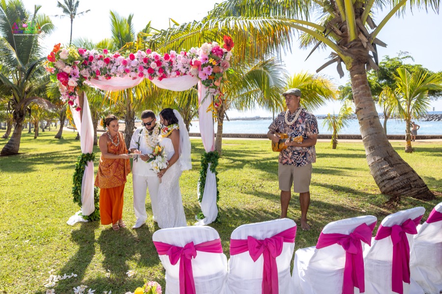 Ukulele player and officiant with the couple under the flower arch, with pink-sashed chairs in front