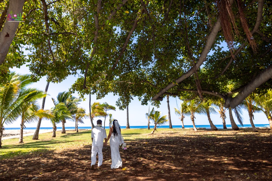 Bride and groom walking hand in hand under shady trees toward the ocean