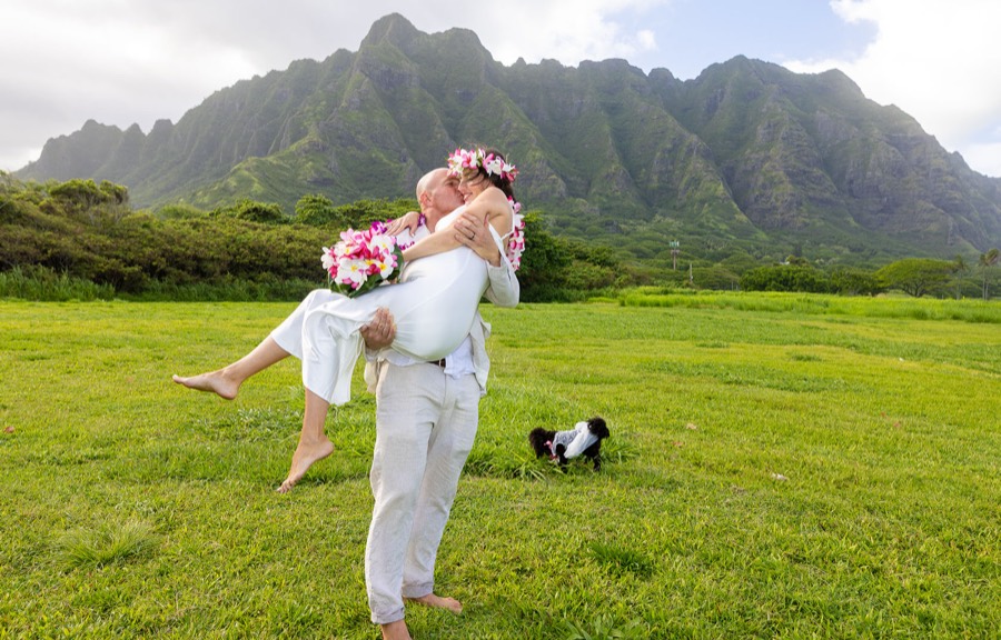 Groom carrying his bride across the grass beneath Kualoa's green mountains, with their little dog beside them