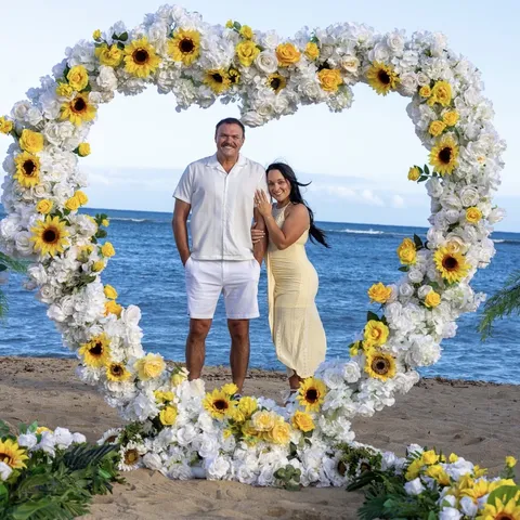 Makenzi and her fianc&eacute; under a heart arch of sunflowers and white flowers on an Oahu beach