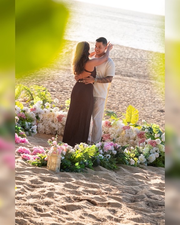 Couple embracing on the sand inside a setup of white and pink roses, tropical leaves and a lantern on an Oahu beach