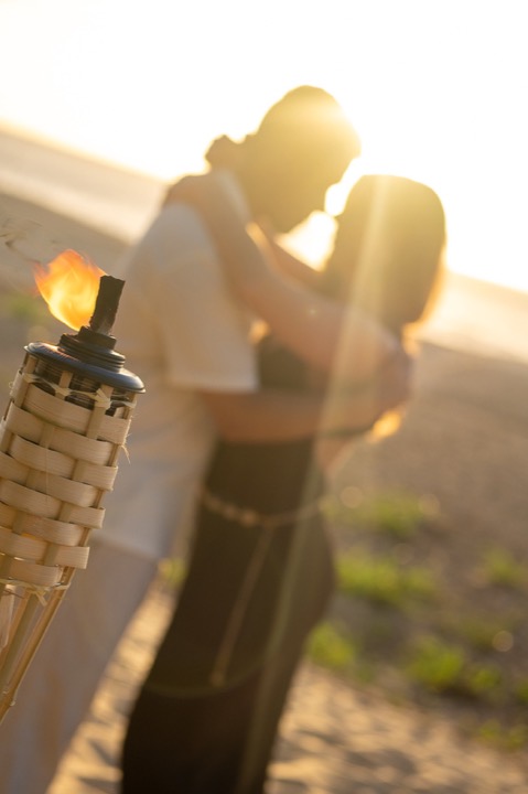 Couple sharing a kiss at sunset on the beach beside a lit bamboo tiki torch