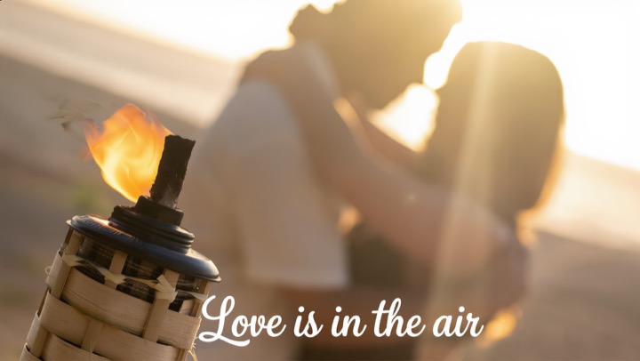 A glowing bamboo tiki torch in front of a couple embracing at golden hour, with the words Love is in the air