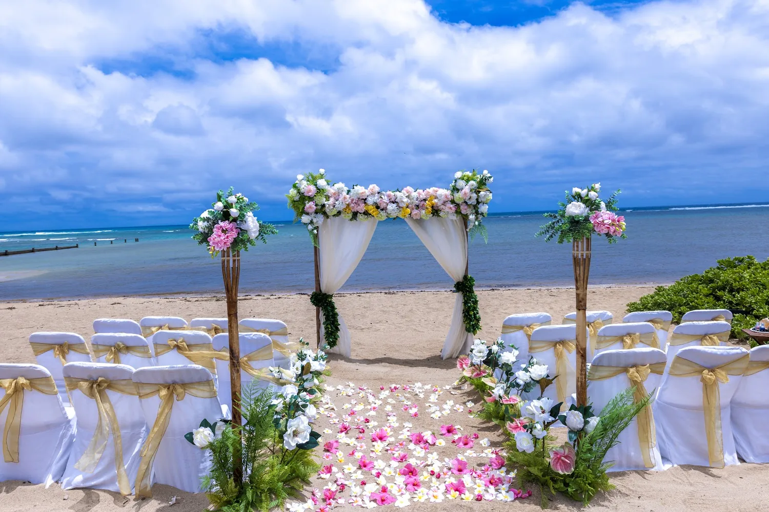 White chairs and a pink and white petal aisle leading to a flower arch on the beach