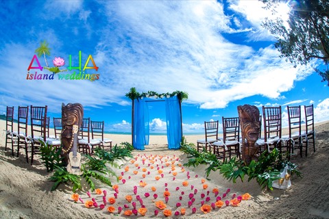 Blue draped bamboo archway with carved tiki men and wooden chairs on the beach at Waimanalo