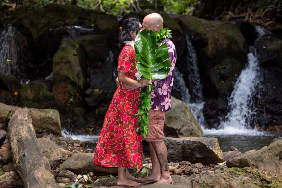 Couple kissing behind a large green leaf in front of a rainforest waterfall on Oahu