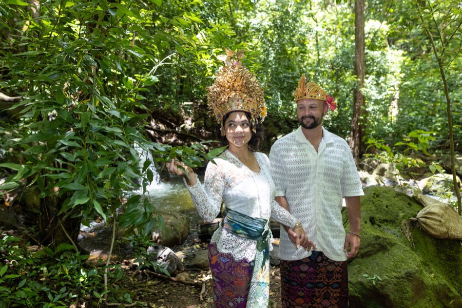 Couple in Balinese-style golden headdresses and traditional sarongs beside a forest waterfall