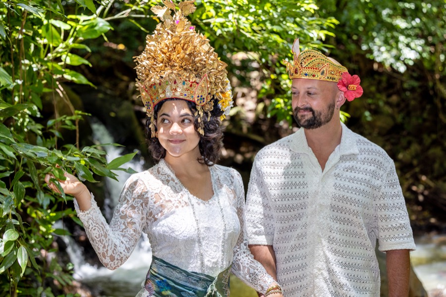 Bride in a golden crown and white lace with her partner among tropical leaves