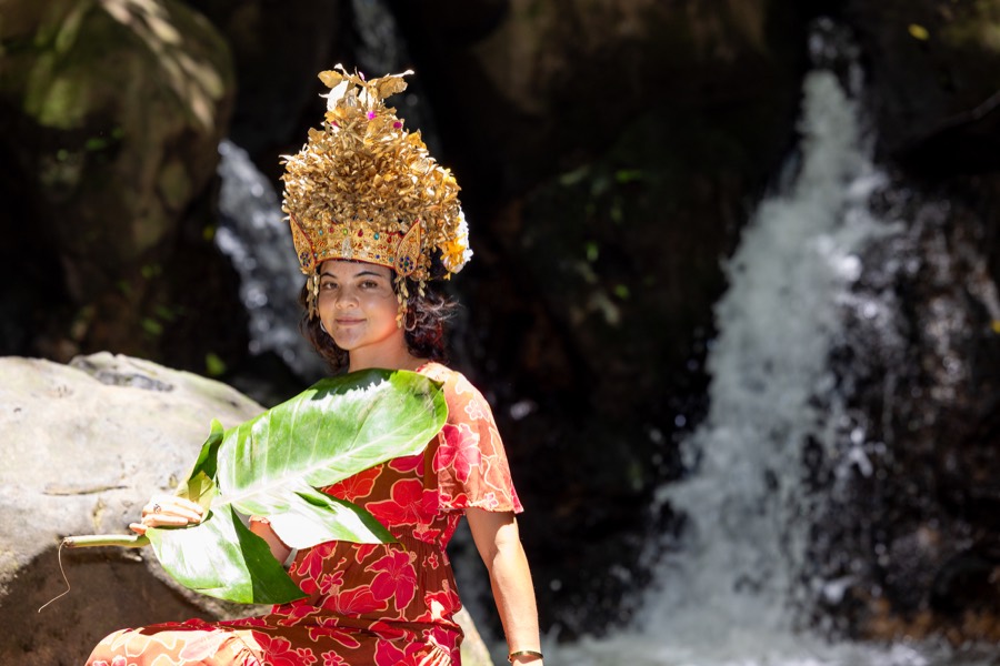 Woman in a golden crown and red dress holding a large leaf in front of a waterfall