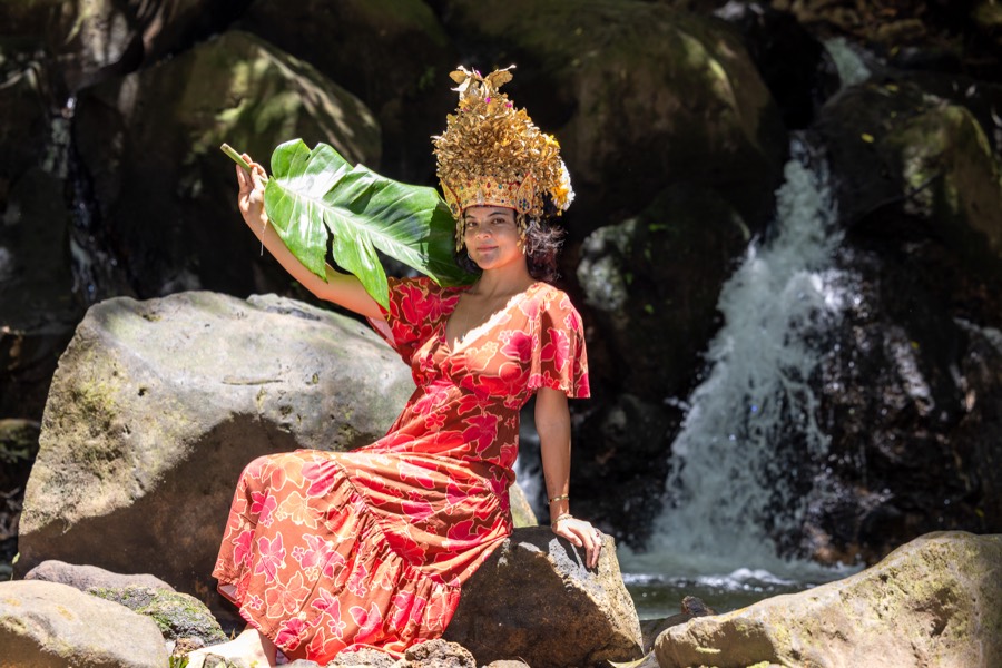 Woman in a golden crown and red floral dress posing on a boulder by a Nuuanu waterfall
