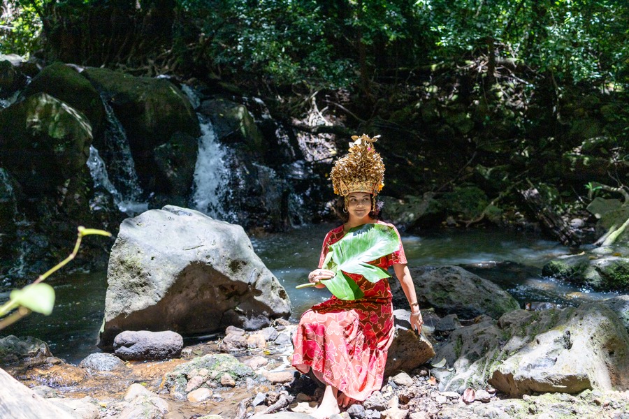 Woman in a red dress and golden crown sitting on the rocks by a waterfall stream