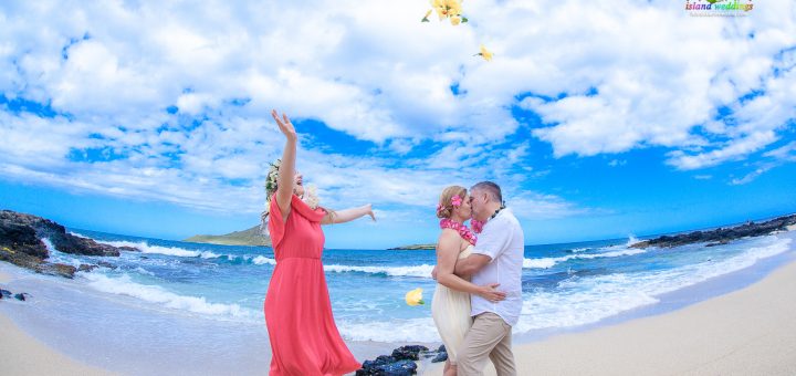 Daughter Emma tosses large yellow hibiscus in the air and her mom and dad kiss on the beach at makapuu
