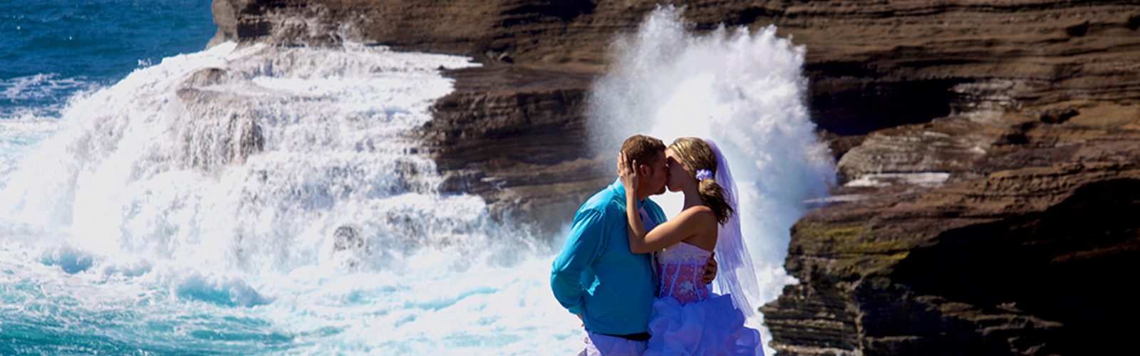 Couple kissing on an ocean cliff as a wave crashes behind them